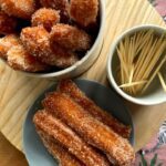 freshly made Churros served in a small bowl and also in a cup with some toothpicks on a sheet