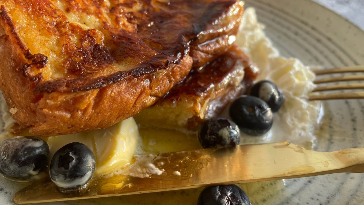 Freshly made 2 french toasts in a plate drizzled with maple syrup and topped with blueberries and whipped cream on a grey plate with fork