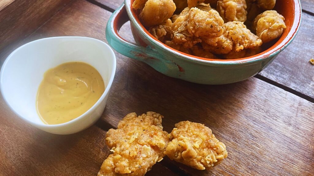 Popcorn chicken served in a blue bowl with dipping sauce on the side on a wooden board