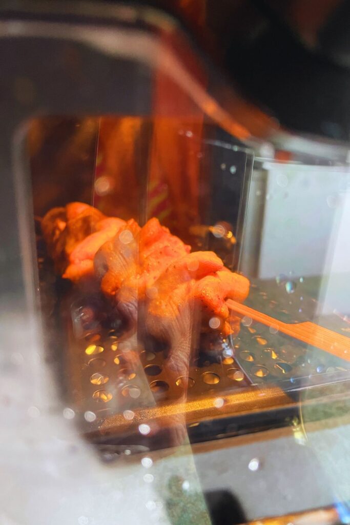 Skewered chicken pieces grilling in the air fryer behind a glass shield, showing visible steam and grill marks, creating an appetizing and warm visual.