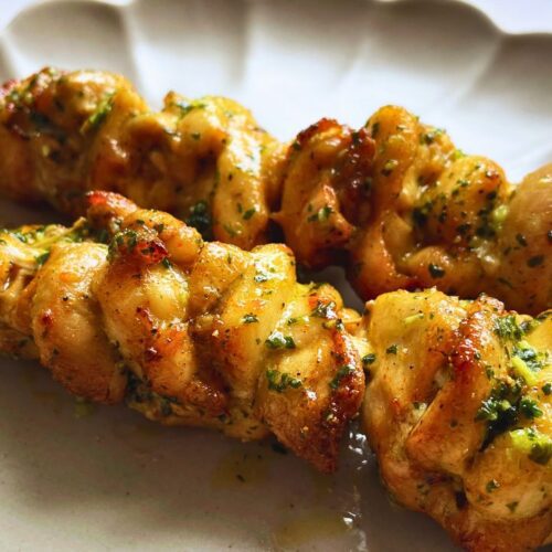 Two grilled chicken skewers garnished with garlic herb butter on a white ceramic plate, displaying a golden-brown color with visible grill marks.