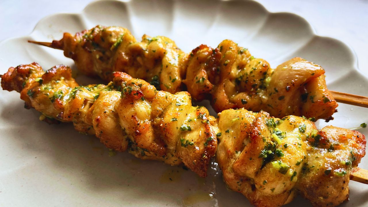 Two grilled chicken skewers garnished with garlic herb butter on a white ceramic plate, displaying a golden-brown color with visible grill marks.