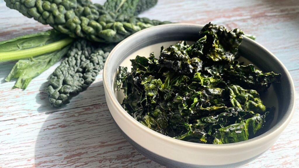 small bowl of kale chips laid on the table with fresh kale leaves on the corner