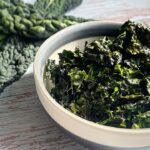 small bowl of kale chips laid on the table with fresh kale leaves on the corner