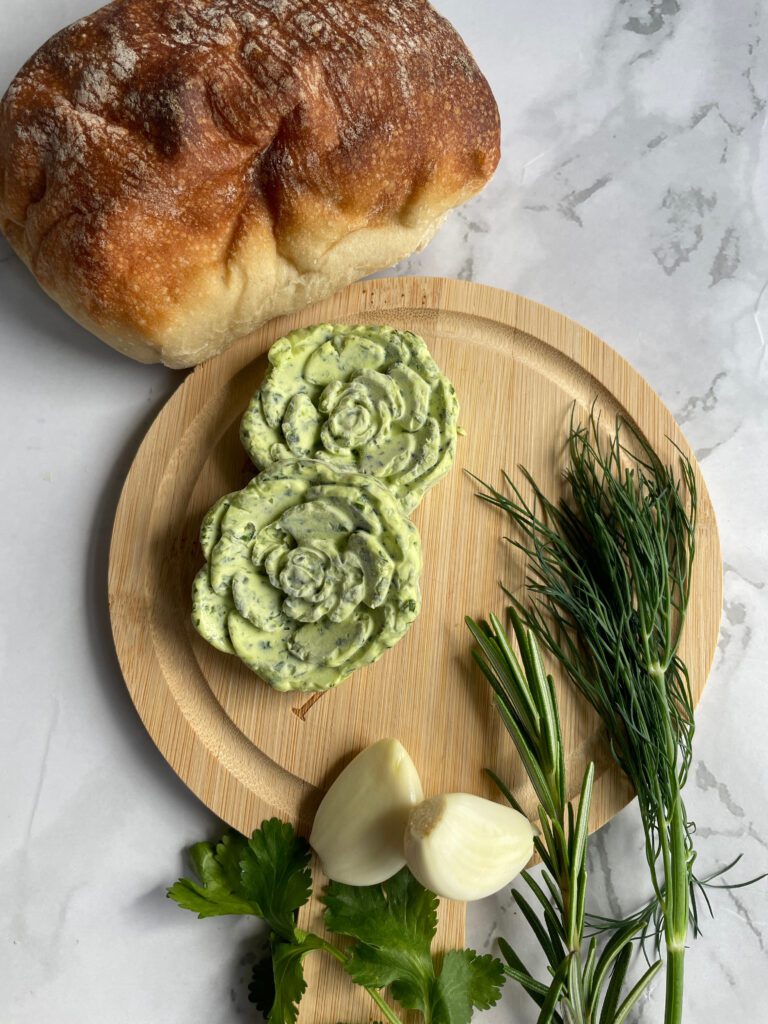 rose shaped herb butter on a round wooden board with fresh herbs and garlic cloves and a sourdough bread
