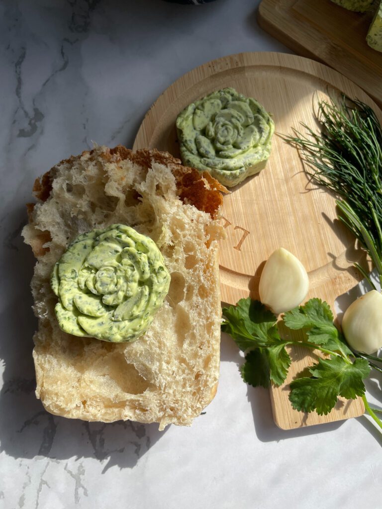 
A wooden cutting board with slices of rustic bread topped with garlic and herb butter. ingredients like garlic cloves, fresh dill, and parsley surround the bread. sunlight illuminates the setup.