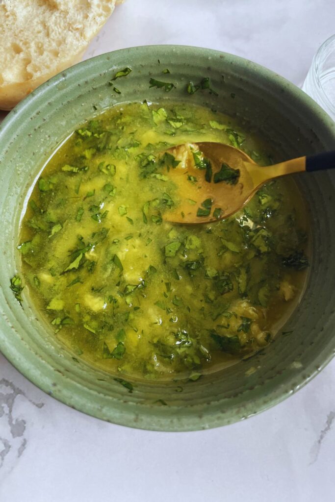 A bowl of vibrant green garlic and herb butter sprinkled with herbs, with a golden spoon, on a white tabletop beside a slice of bread.