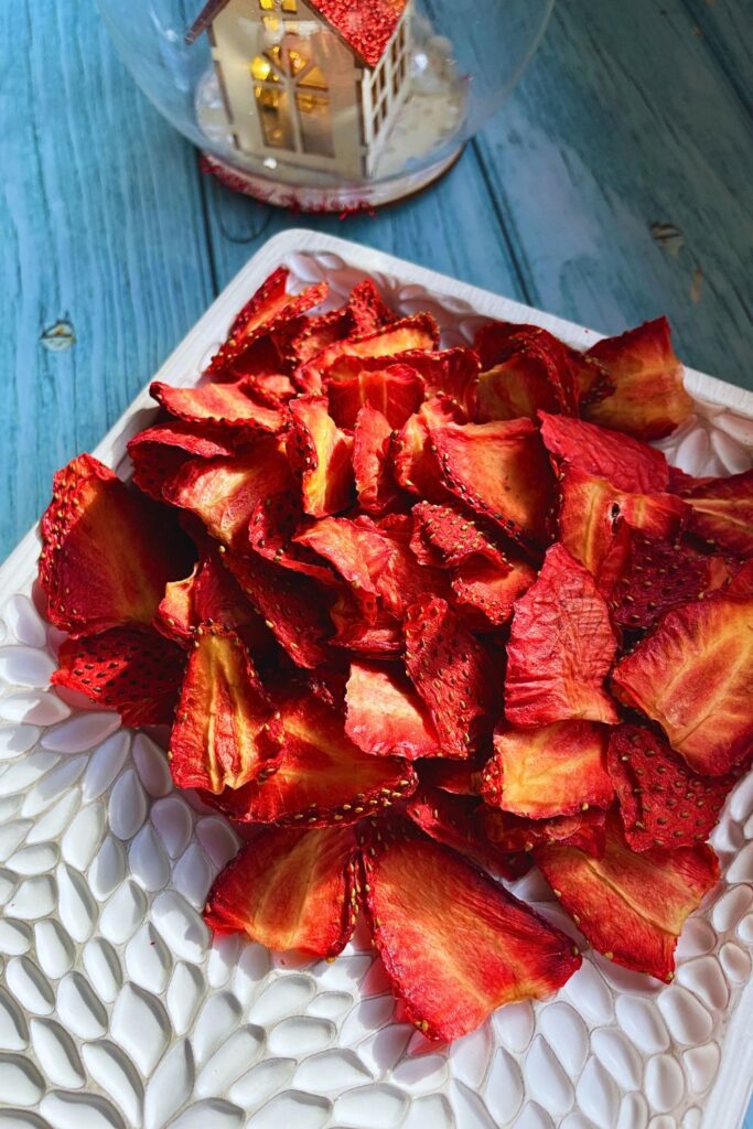 A pile of air fryer dried strawberry slices is displayed on a white decorative plate. The strawberries are vibrant red and textured. In the background, there is a glass dome containing a small house with a red roof, adding a charming touch to the scene.