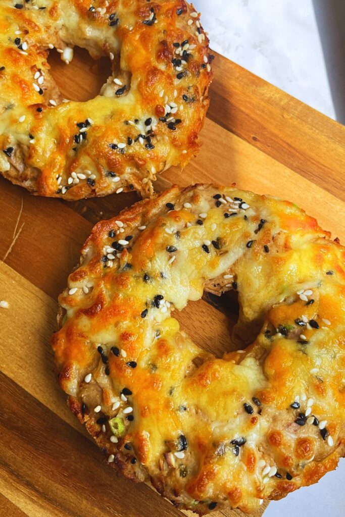 A close-up image of two freshly baked Air fryer tuna melt on bagels topped with melted cheese and sprinkled with sesame seeds. The bagels are placed on a wooden surface, highlighting their crispy, golden-brown texture and savory toppings.