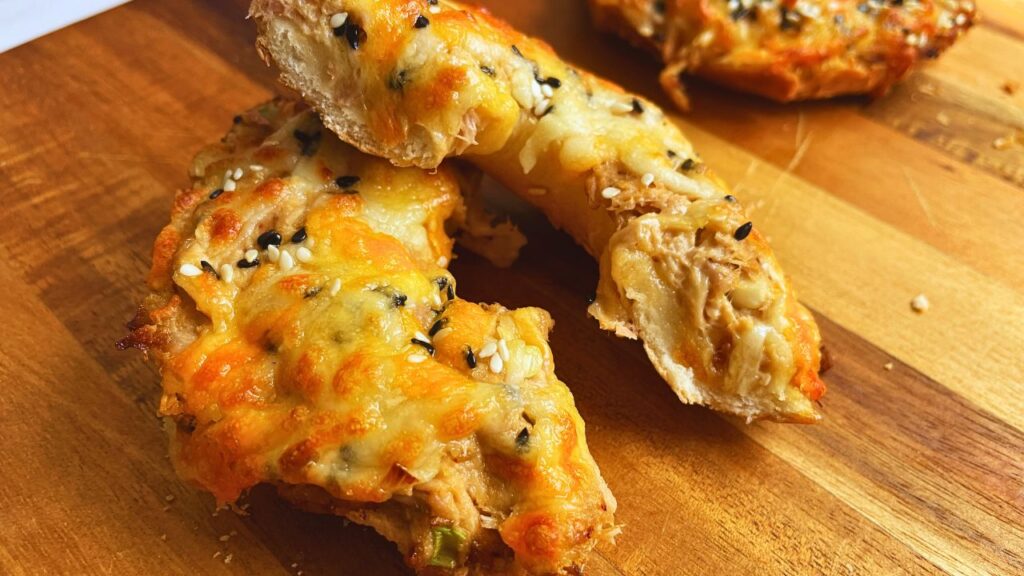 Freshly baked tuna melt on bagels cut in halves. The bagels are placed on a wooden surface, highlighting their crispy, golden-brown texture and savory toppings.