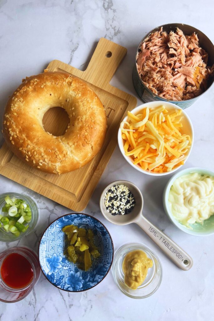 Air fryer tuna melt on bagel ingredients list; bagel, cheese, mayonnaise, mustard, siracha sauce, spring onions, bagel seasoning, jalapeno laid on a marble kitchen counter.