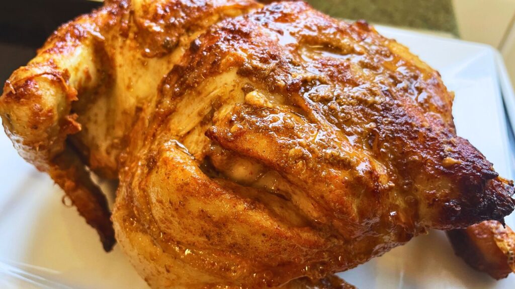 A close-up image of a golden-brown air fryer roasted chicken with crispy skin, served on a white plate. the chicken appears juicy and well-seasoned.