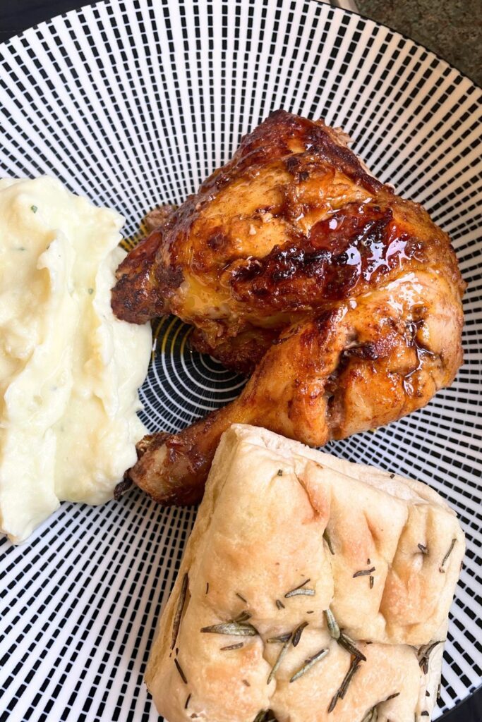 A plate with a grilled air frier chicken leg, mashed potatoes, and a piece of rosemary focaccia bread, viewed from above. the plate has a black and white pattern.