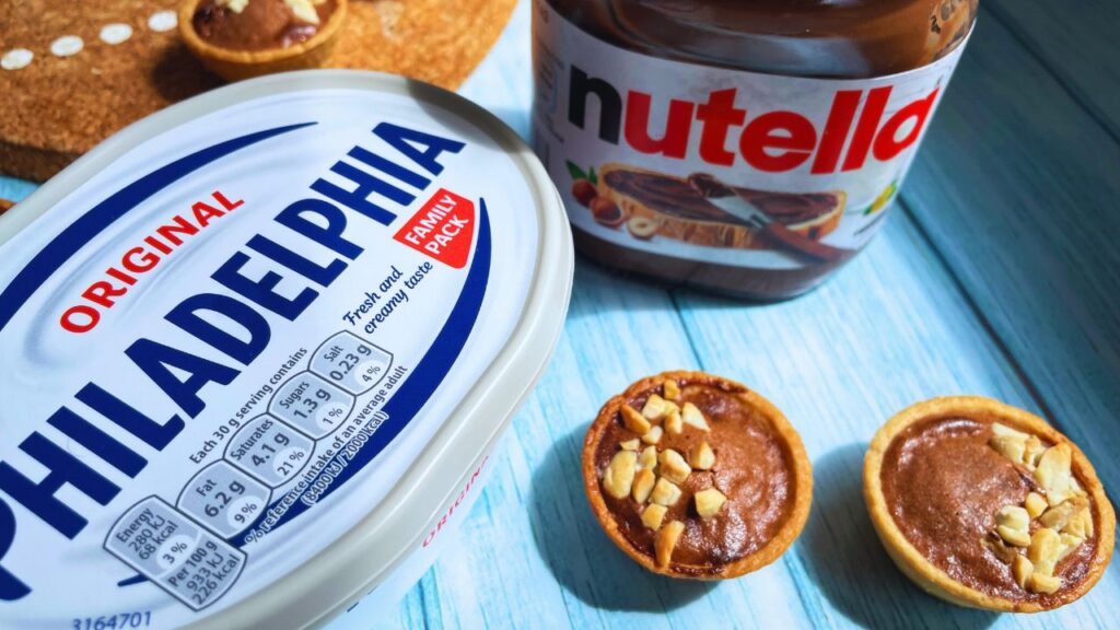 Mini air fryer Nutella tarts on a blue wooden table with Nutella jar on the back and a tub of Philadelphia cream cheese.