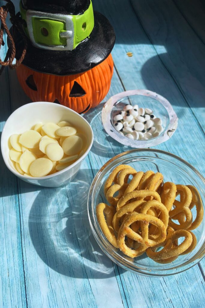 Halloween Monster Pretzels Ingredients Laid In A Blue Board Pretzels, White Chocolate Melts, Googly Candy Eyes