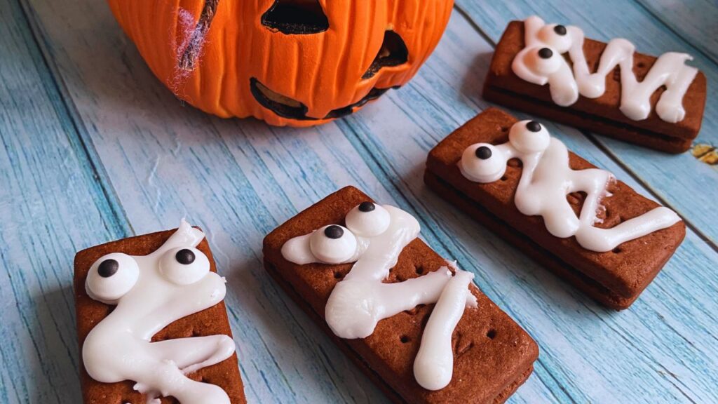 Four rectangular Burboun biscuits are decorated to look like mummies with white icing and candy googly eyes. They are arranged on a bright blue wooden board with a Halloween pumpkin at the back.surface.Halloween Mummy Biscuits